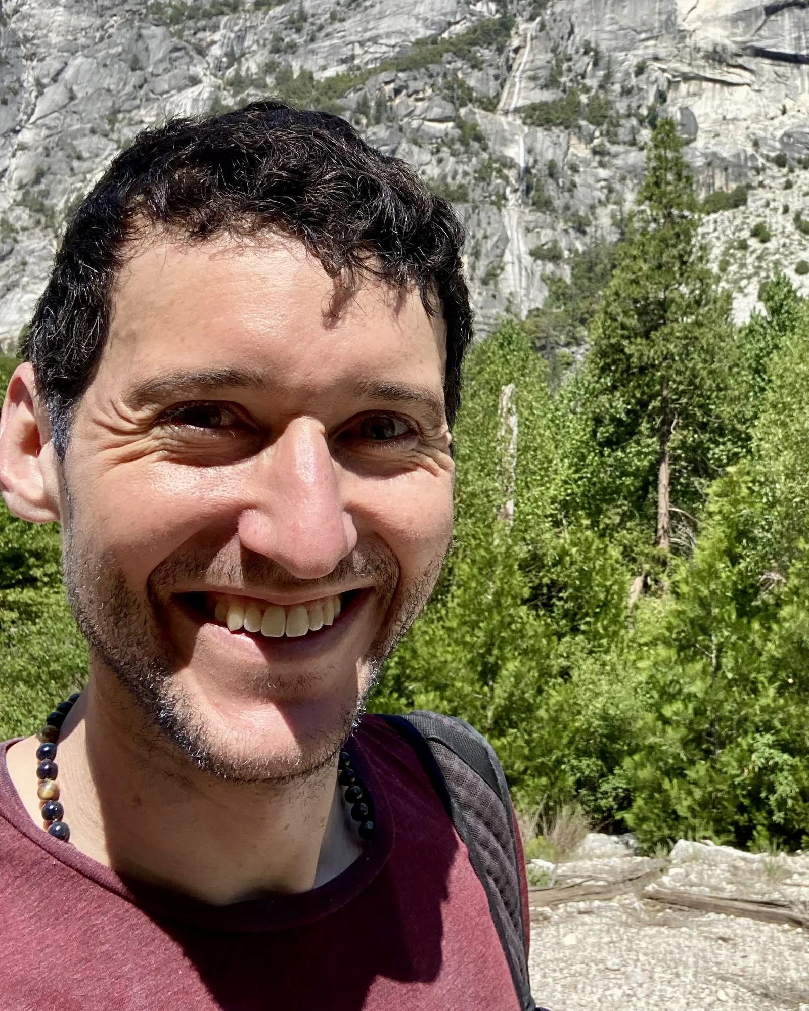 Greg outdoors, smiling at the camera in close-up, with a granite cliff face and conifers behind him.
